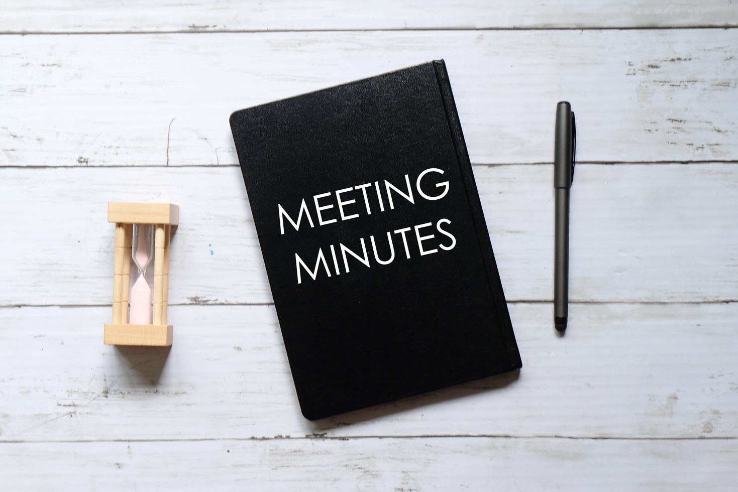 Top view of hour glass,pen and notebook written with 'MEETING MINUTES' on white wooden background.